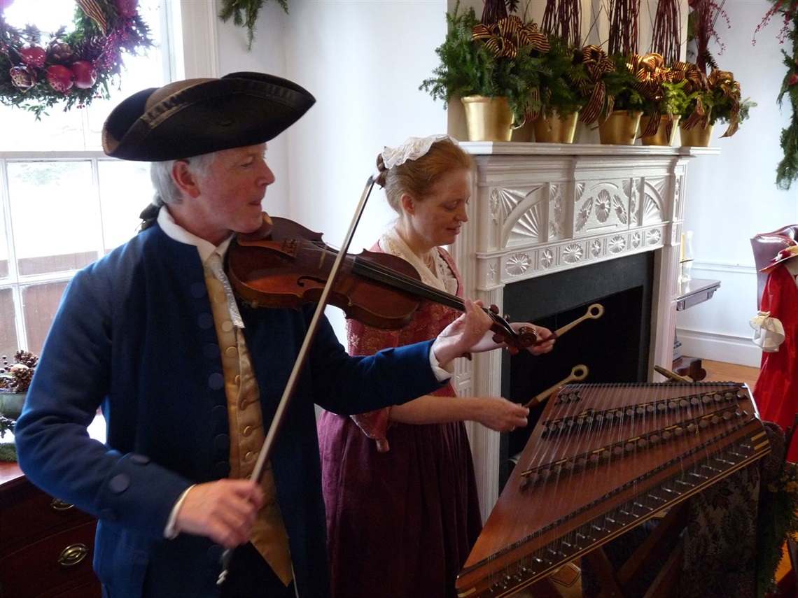 Anne and Ridley Enslow in colonial period dress inside a period sitting room. Ridley is playing violin and Anne is playing hammered dulcimar.