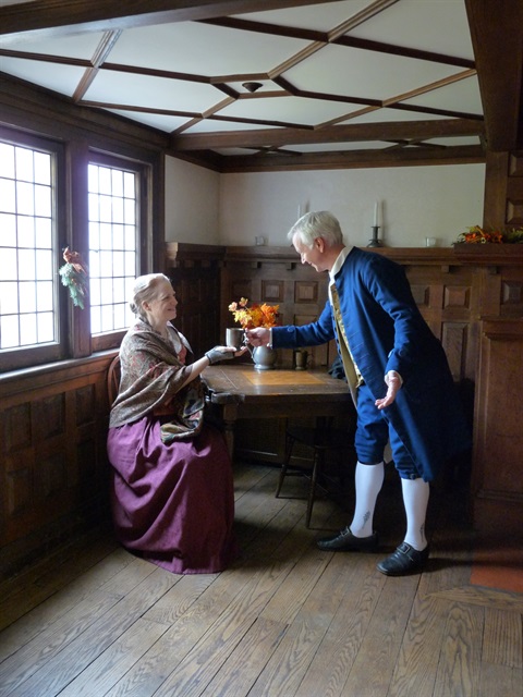 Historical entertainers Anne and Ridley Enslow in colonial dress inside a colonial home or tavern, sharing a toast.